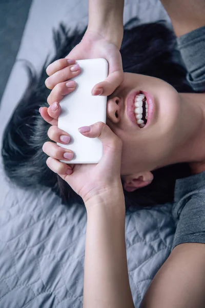 Mujer deprimida en la cama cubriendo la cara con teléfono inteligente en el dormitorio - foto de stock
