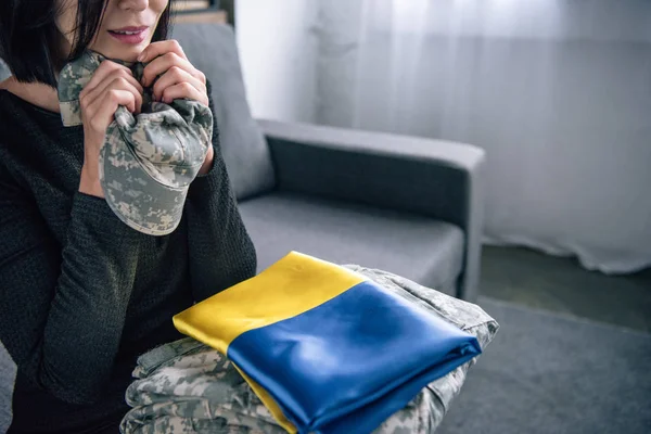 Обрезанный вид расстроенной женщины, держащей военную одежду и украинский флаг дома — стоковое фото