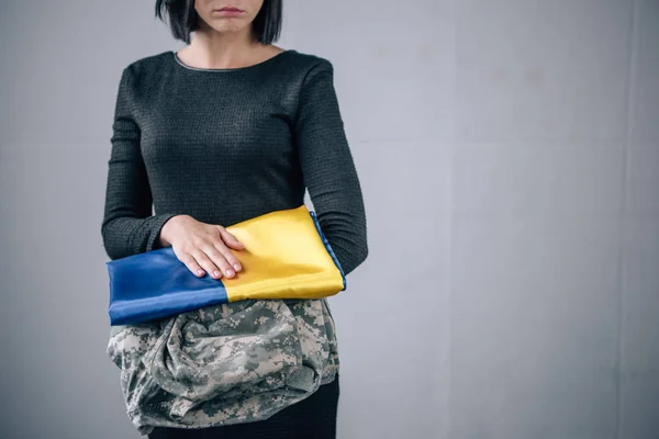 Partial view of upset woman holding military clothing and ukrainian flag at home — Stock Photo