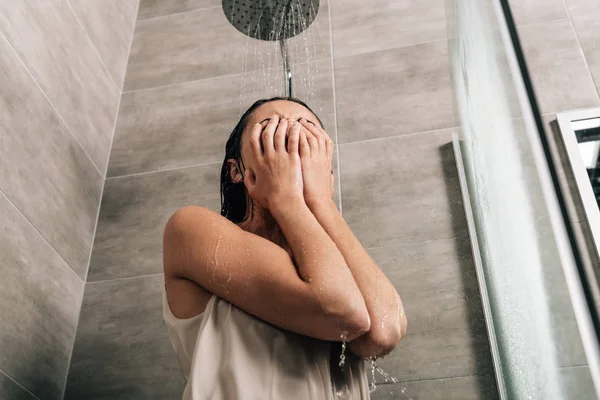 Mujer deprimida cubriendo la cara con las manos mientras llora en la ducha en casa - foto de stock