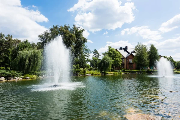Fountains in pond near trees and house in summertime — Stock Photo