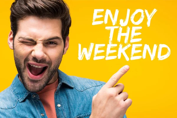 Handsome man pointing with finger at enjoy the weekend illustration and winking isolated on yellow — Stock Photo