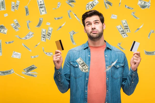Confused handsome man holding credit cards isolated on yellow with money rain illustration — Stock Photo