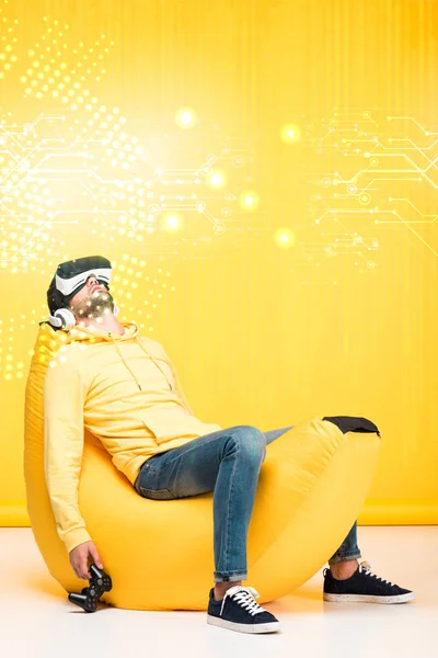 KYIV, UCRANIA - 12 DE ABRIL: hombre durmiendo en la silla de la bolsa de frijoles con joystick en auriculares de realidad virtual en amarillo con ilustración del ciberespacio - foto de stock