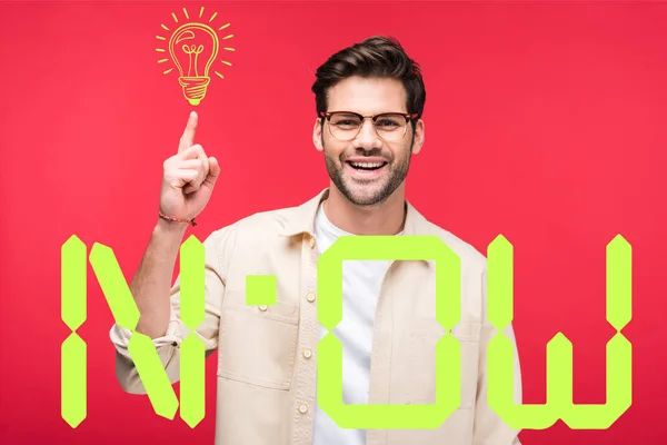 Uomo bello sorridente che punta con il dito alla lampadina isolata su rosso con ora lettering — Foto stock