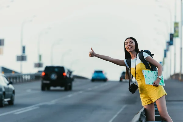 Belle et asiatique femme en salopettes jaunes auto-stop et tenant carte — Photo de stock