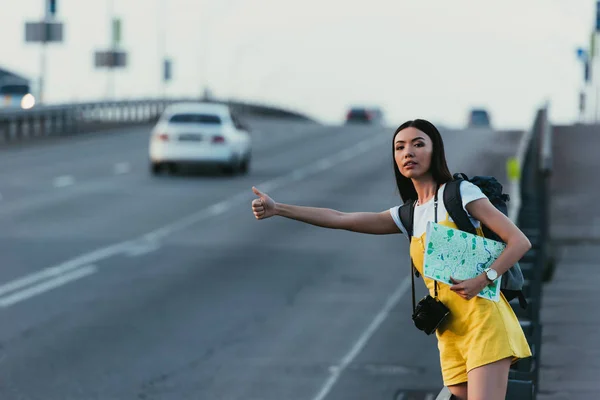 Asiático mujer en amarillo overoles autoestop y celebración de mapa - foto de stock