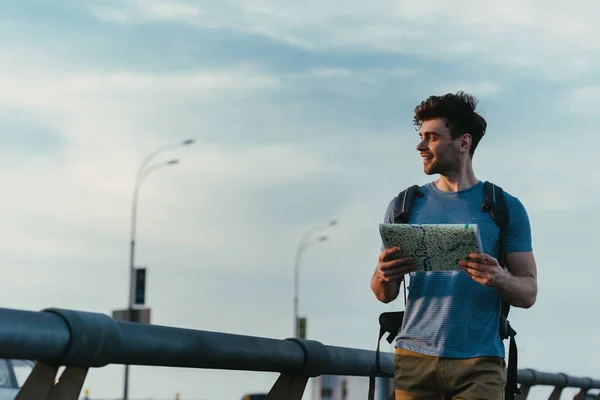 Uomo sorridente e bello in t-shirt e pantaloncini che tiene la mappa e distoglie lo sguardo — Foto stock