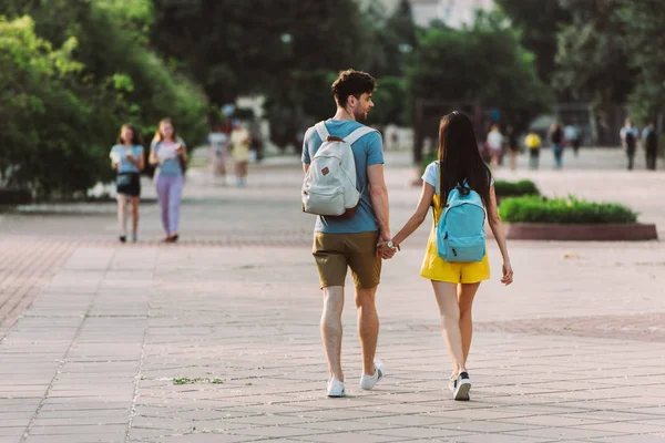 Back view of man and woman with backpacks walking and holding hands — Stock Photo