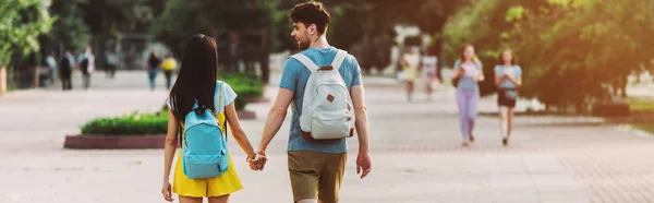 Panoramic shot of man and woman with backpacks walking and holding hands — Stock Photo