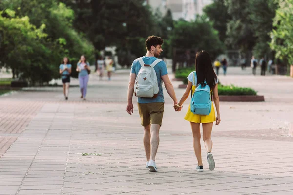 Vista trasera del hombre y la mujer con mochilas caminando y tomados de la mano - foto de stock