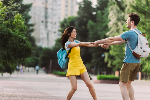 Guapo hombre y asiático mujer con mochilas sonriendo y cogido de la mano - foto de stock