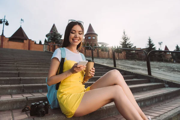 Улыбающаяся и азиатская женщина, использующая смартфон и держащая бумажную чашку — Stock Photo