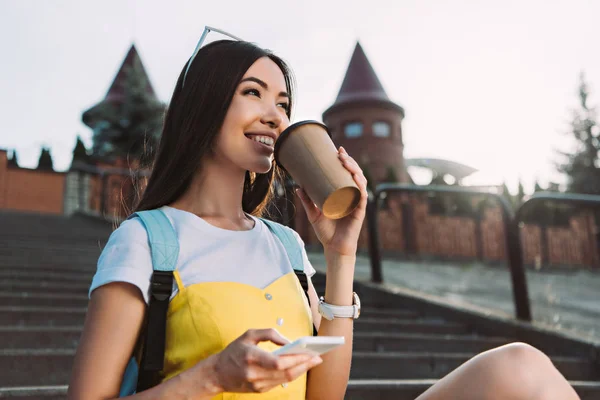 Sonriente y asiático mujer sosteniendo smartphone y bebiendo café - foto de stock