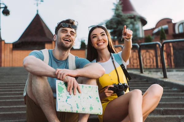 Sorridente e asiatica donna che punta con dito e bell'uomo che tiene la mappa — Foto stock