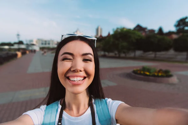 Atractiva y asiática mujer con gafas sonriendo y tomando selfie - foto de stock