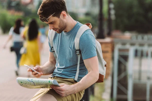 Hombre guapo en camiseta usando smartphone y sosteniendo mapa - foto de stock