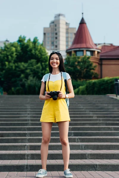 Atractiva y asiática mujer en overoles sosteniendo cámara digital y mirando a la cámara - foto de stock