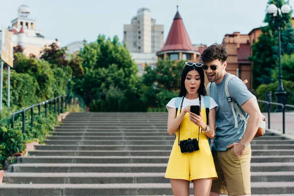 Sonriente y guapo hombre y asiático mujer mirando smartphone - foto de stock