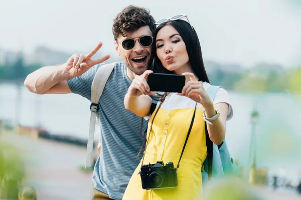Guapo hombre mostrando paz signo y asiático mujer tomando selfie - foto de stock