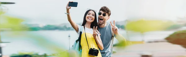 Tiro panorámico de hombre guapo y mujer asiática mostrando signos de paz y tomando selfie - foto de stock