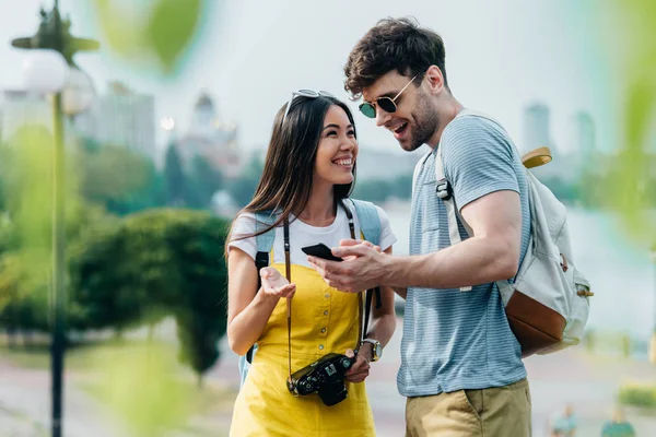 Guapo hombre sosteniendo smartphone y asiático mujer hablando con él - foto de stock