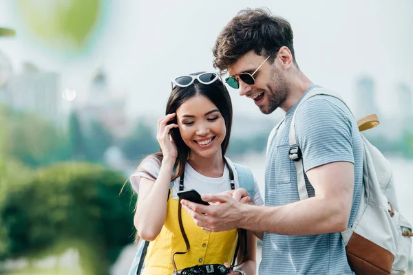 Guapo hombre y asiático mujer sonriendo y mirando el teléfono inteligente - foto de stock