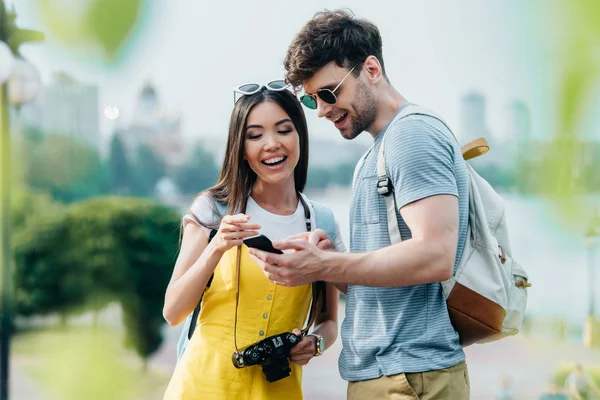 Guapo hombre y asiático mujer sonriendo y mirando el teléfono inteligente - foto de stock