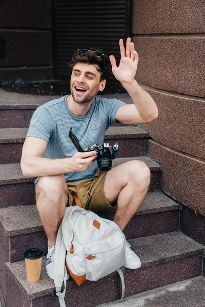 Sonriente y guapo hombre en gafas ondeando y sosteniendo cámara digital - foto de stock