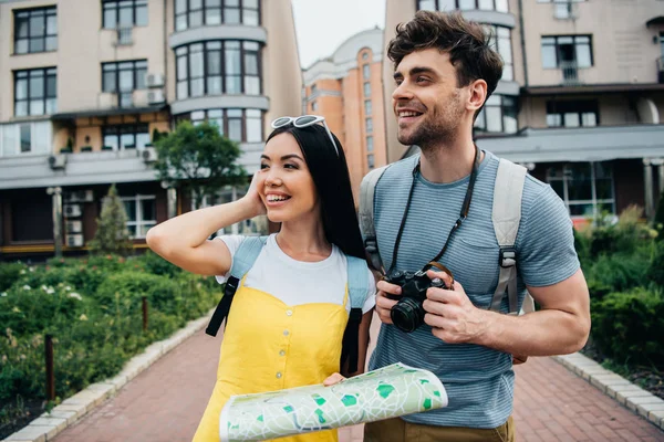 Hombre guapo con cámara digital y mujer asiática con mapa mirando hacia otro lado - foto de stock