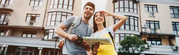 Panoramic shot of handsome man with digital camera and asian woman with map looking away — Stock Photo