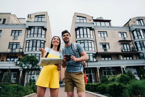 Bel homme avec appareil photo numérique et femme asiatique avec carte regardant loin — Photo de stock