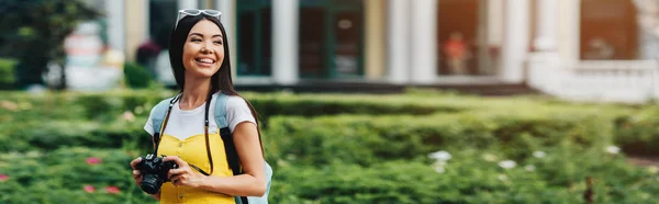 Tiro panorámico de mujer atractiva y asiática con cámara digital mirando hacia otro lado - foto de stock