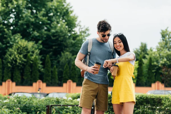 Hombre guapo y mujer asiática mirando reloj y sosteniendo vasos de papel - foto de stock