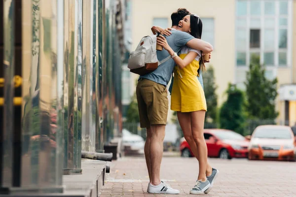 Hombre con mochila y mujer asiática abrazando y sosteniendo vasos de papel - foto de stock