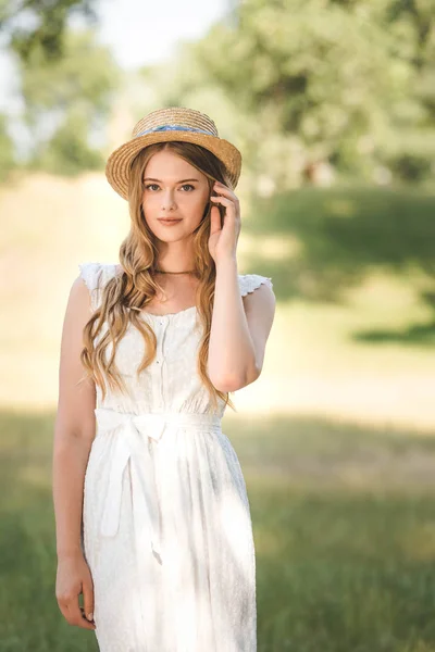 Hermosa chica con sombrero de paja de pie en el prado y mirando a la cámara - foto de stock