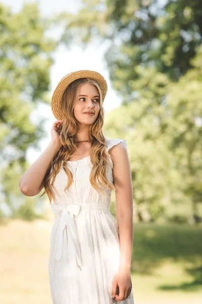 Hermosa chica con sombrero de paja de pie en el prado y mirando hacia otro lado - foto de stock