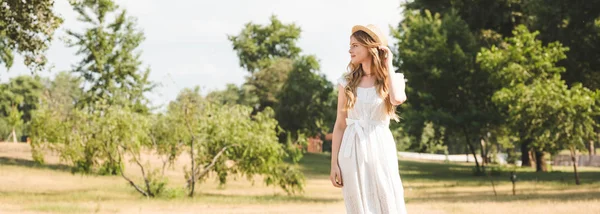 Panoramic shot of beautiful girl with straw hat and white dress standing on meadow and looking away — Stock Photo