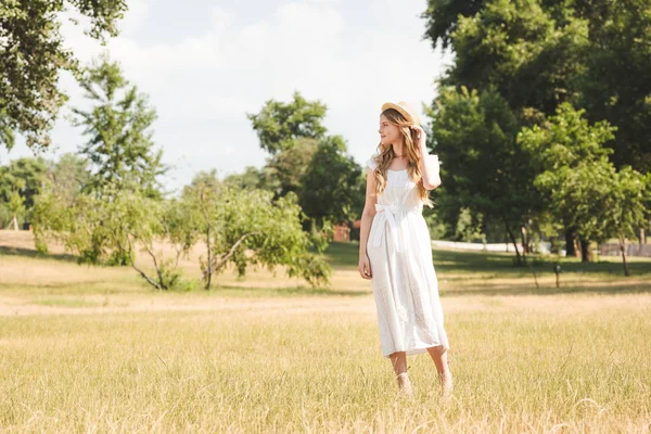 Beautiful girl in white dress and straw hat standing on meadow and looking away — Stock Photo