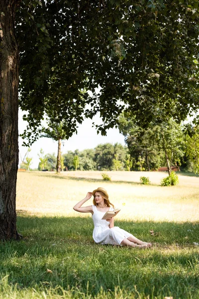 Full length view of beautiful girl in white dress touching straw hat and holding book while sitting on meadow and looking away — Stock Photo