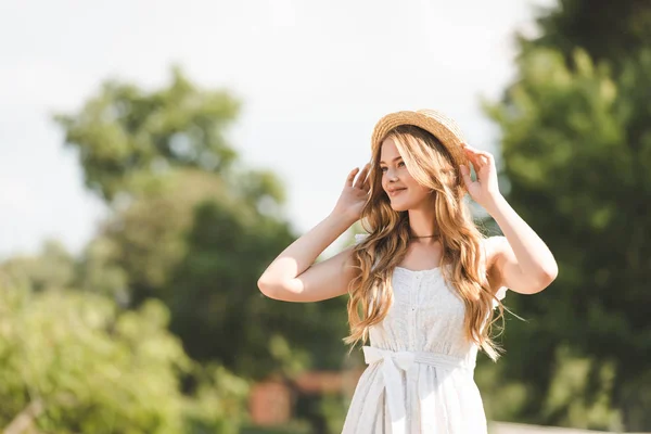 Hermosa chica en vestido blanco tocando sombrero de paja y sonriendo mientras está de pie en el prado y mirando hacia otro lado - foto de stock
