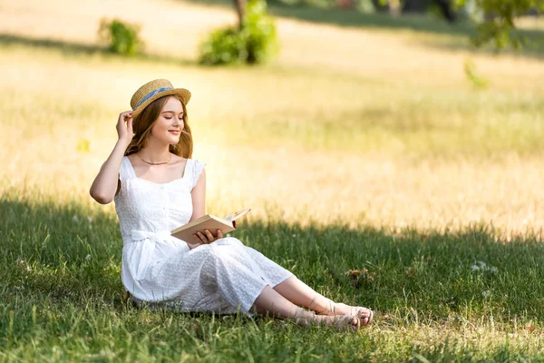 Schönes Mädchen in weißem Kleid, das Strohhut berührt und Buch hält, während es mit geschlossenen Augen auf der Wiese sitzt — Stockfoto