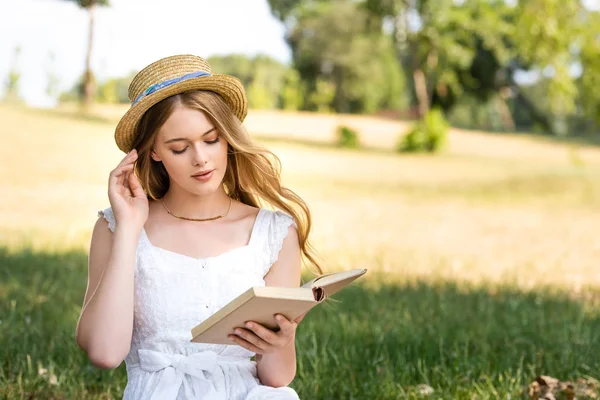 Beautiful girl in white dress touching straw hat and reading book while sitting on meadow — Stock Photo