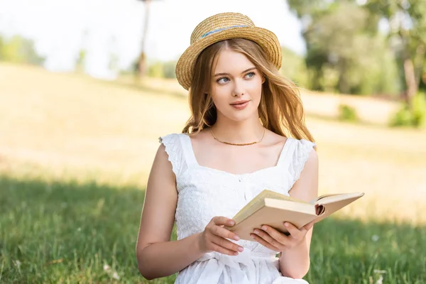 Beautiful girl in white dress and straw hat holding book while sitting on meadow and looking away — Stock Photo