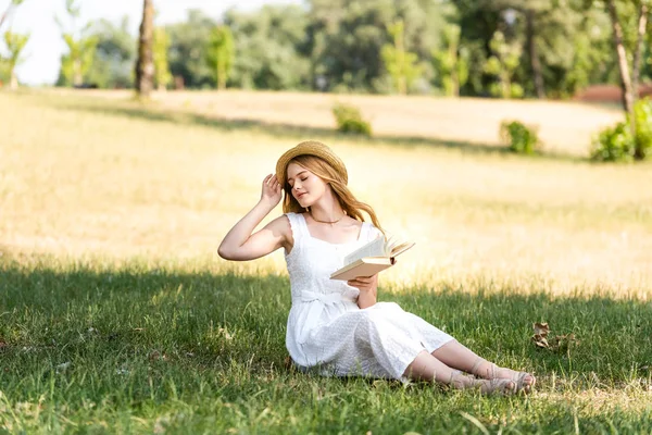 Visão de comprimento total da menina bonita em vestido branco tocando chapéu de palha e segurando livro enquanto sentado no prado com olhos fechados — Fotografia de Stock