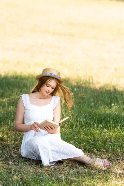 Full length view of beautiful girl in white dress and straw hat reading book while sitting on meadow — Stock Photo