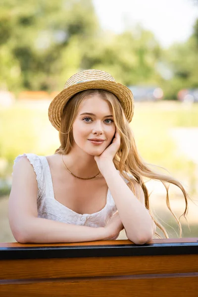 Schönes Mädchen mit Strohhut posiert auf Bank sitzend und in die Kamera blickend — Stockfoto