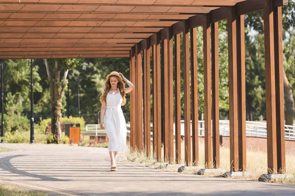 Vue pleine longueur de belle fille en robe blanche et chapeau de paille marchant près de la construction en bois — Photo de stock