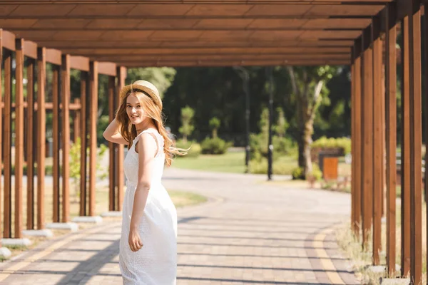 Красивая девушка в белом платье касаясь соломенной шляпы во время прогулки возле деревянной конструкции и глядя на камеру — стоковое фото