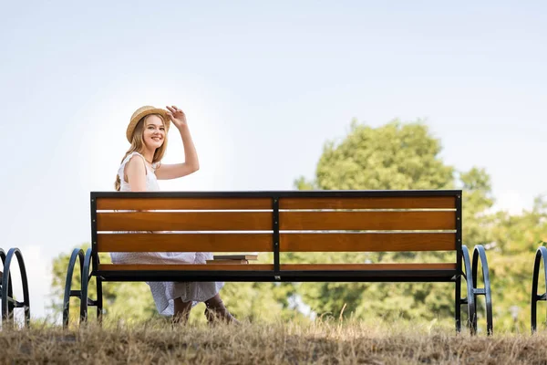 Schönes Mädchen in weißem Kleid, das Strohhut berührt und lächelt, während es auf der Bank sitzt und in die Kamera schaut — Stockfoto